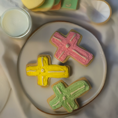 cross cut out sugar cookie on a white cloth background, decorated with yellow green and pink buttercream frosting, beside a cup of milk.