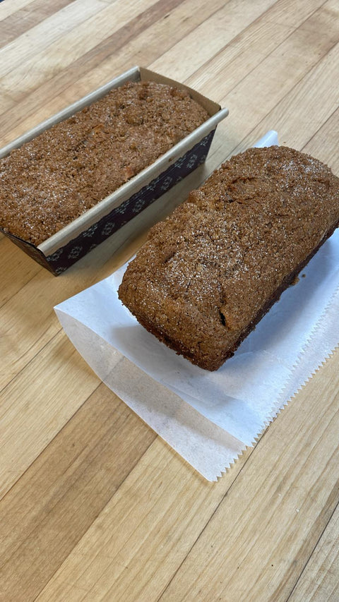 A paper-wrapped apple cinnamon bread loaf beside a smooth, golden brown loaf on a wooden board